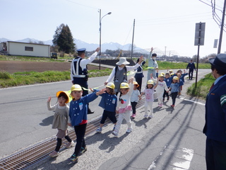 4歳児が駐在さんに見守られ、手を挙げて道を渡っています
